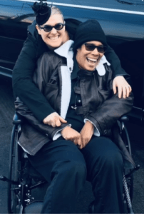 Larissa is seated in a black wheelchair with her arms in the center. She is wearing all black. She has a bright smile. Her partner Shayna embraces her from behind also wearing all black and wearing a bright smile. 